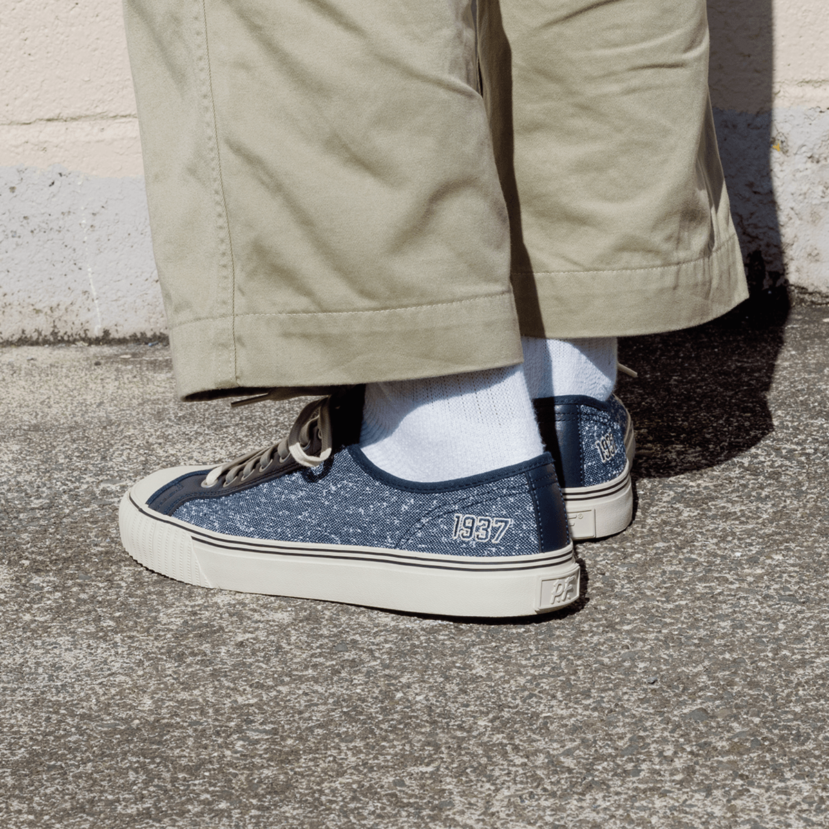 Navy jersey-textured low-top sneakers stamped ‘1937,’ worn with khaki pants, standing against a concrete wall.