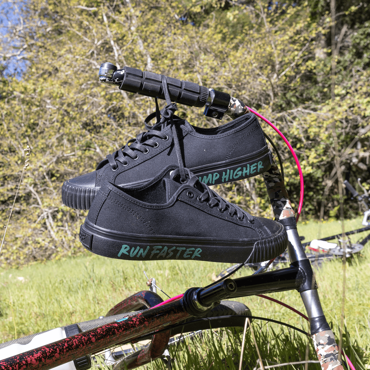 A close-up of black low-top sneakers hanging from a bike handle.