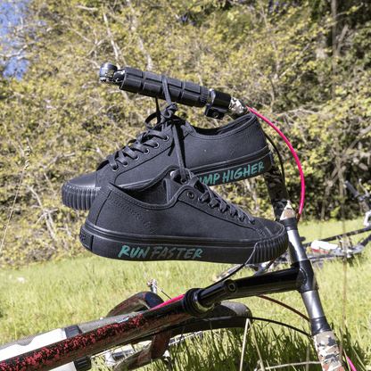 A close-up of black low-top sneakers hanging from a bike handle.