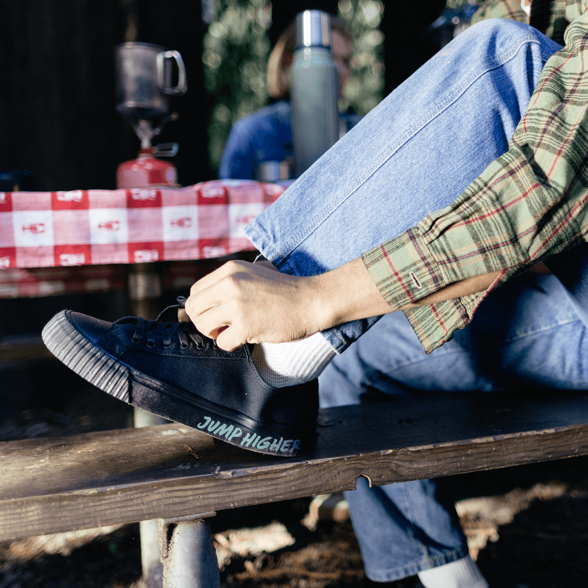 Person sitting on a bench wearing black low-top shoes, blue jeans, and a plaid shirt, with a picnic setup in the background.