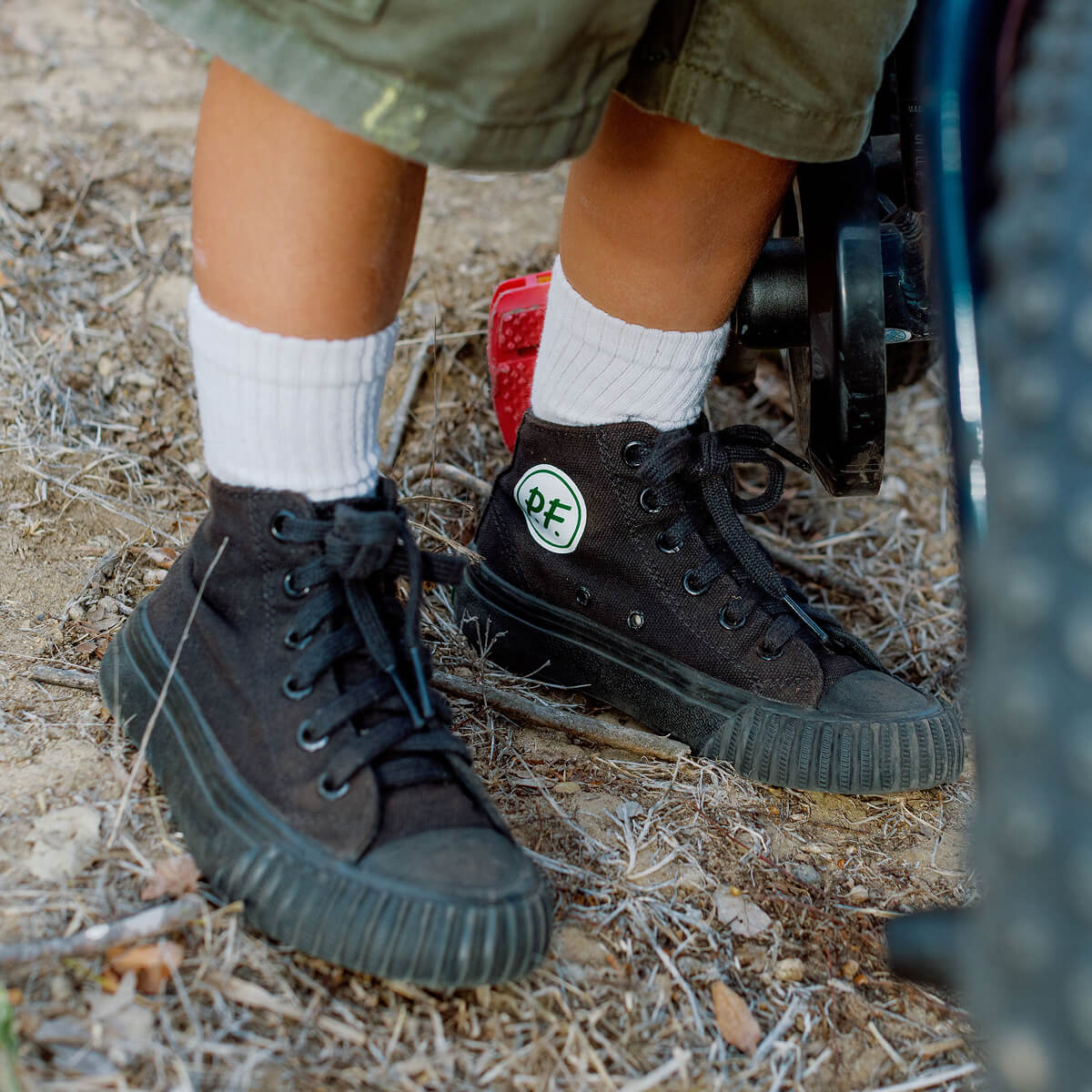 Close-up of a child wearing black canvas high-top sneakers, standing on dry grass near a bicycle.