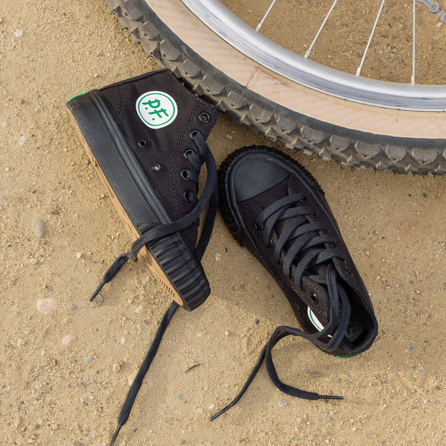 A pair of kids' black high-top sneakers with textured soles, placed on sandy ground next to a bicycle tire.