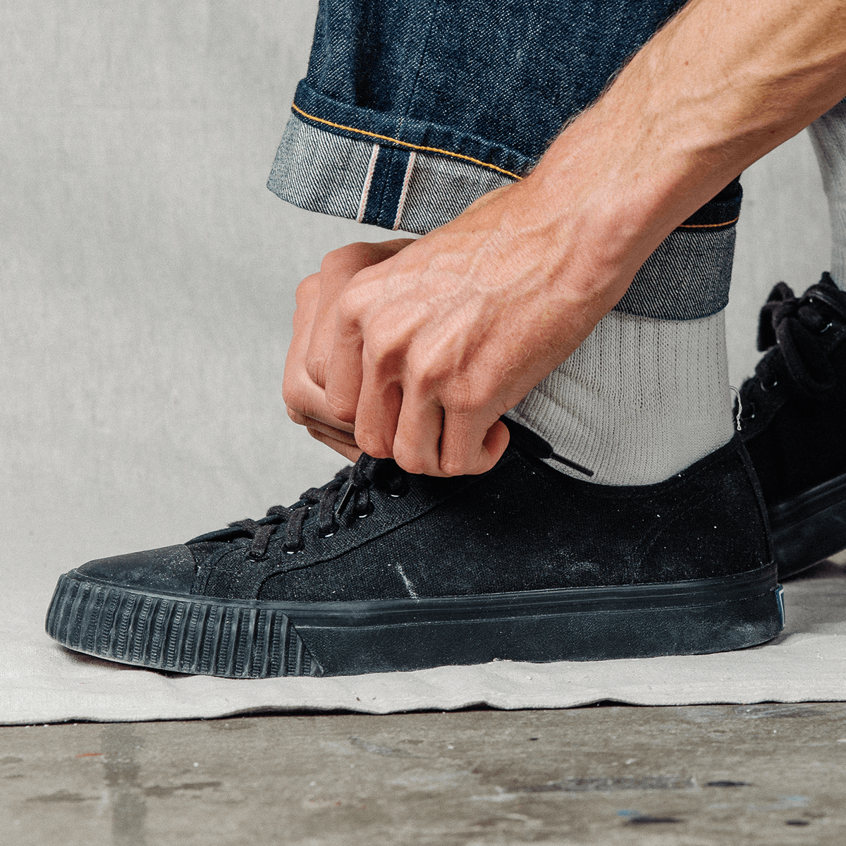 Person tying the laces of a black low-top sneaker on a neutral background.
