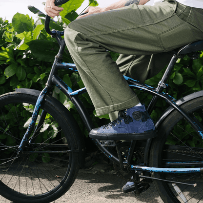 Person riding a bicycle with green pants and blue shoes on a path surrounded by greenery.