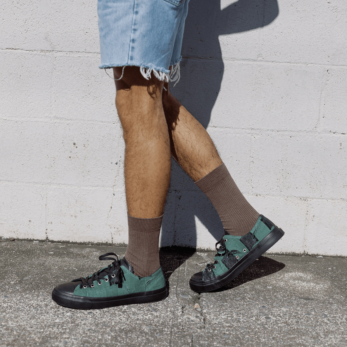 Person wearing green sneakers and brown socks against a white wall.