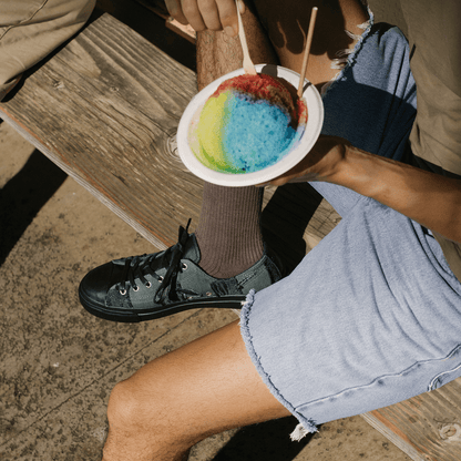 Person holding a colorful shave ice dessert on a wooden bench wearing P.F. Flyers sneakers.
