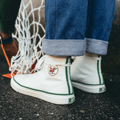 White high-top sneakers with green accents worn by a person standing next to a basketball hoop net.
