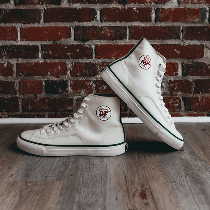 White high-top sneakers with green accents on a wooden floor with a brick wall background.