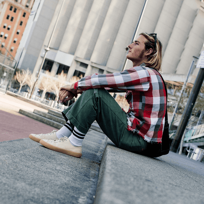 Person sitting on a concrete ledge wearing a plaid shirt, green pants, and beige low-top sneakers in an urban setting.