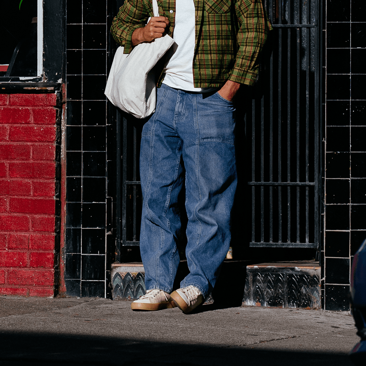 Person wearing a plaid shirt, blue jeans, and beige sneakers standing against a black tiled wall.