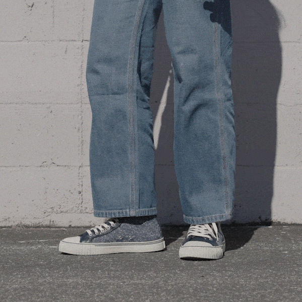 Person wearing navy low-top sneakers and blue jeans shifting stance against a stone wall.