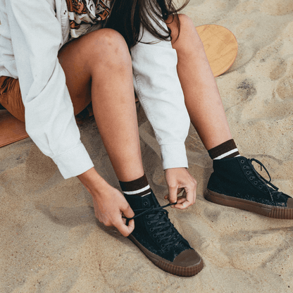 Person seated on a sandboard, tightening the laces of black high-top sneakers.