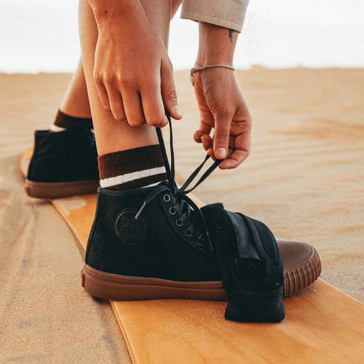 Person tying the laces of a black high-top shoe while standing on a sandboard.