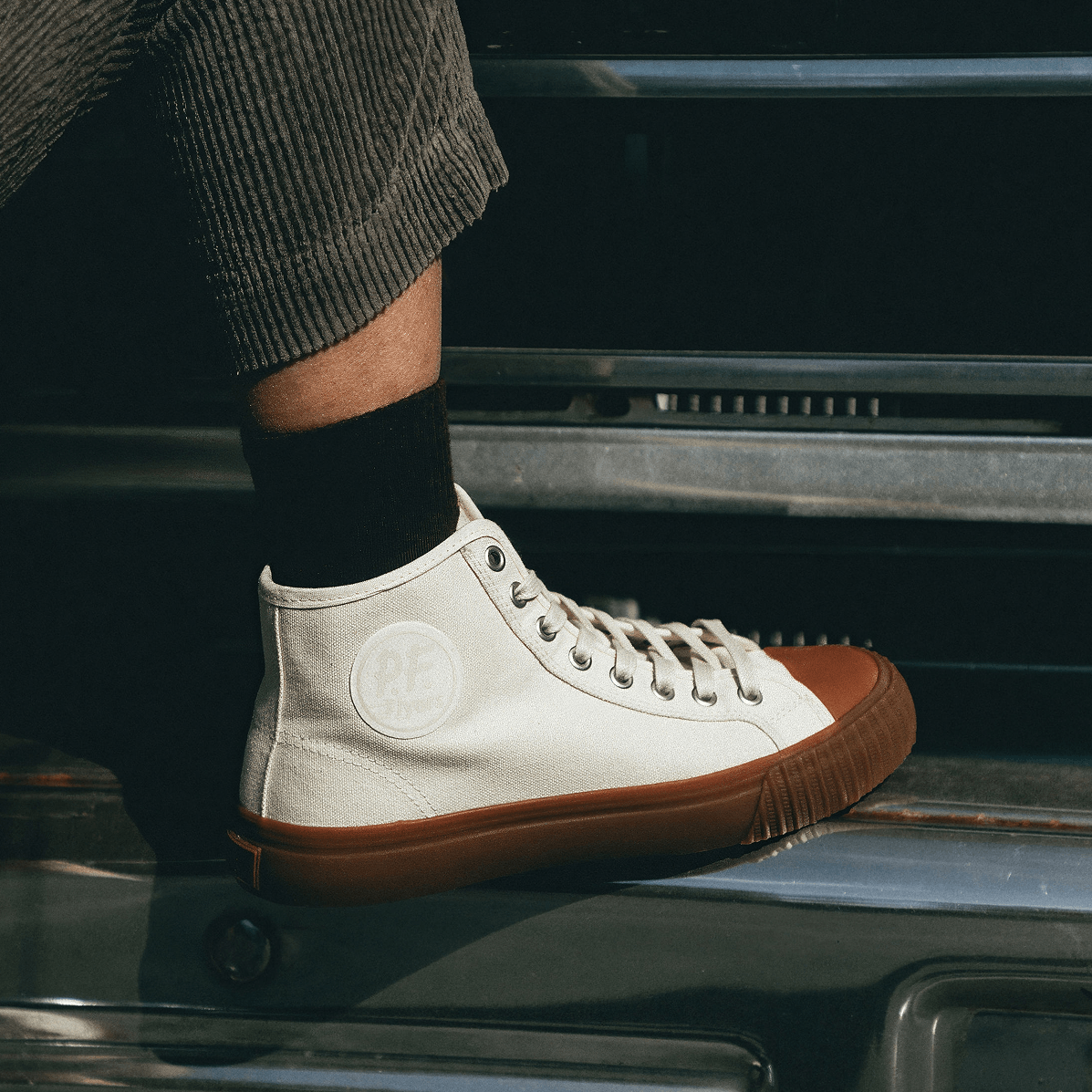 Person wearing a white high-top sneaker with a brown sole, resting their foot on a truck bumper.
