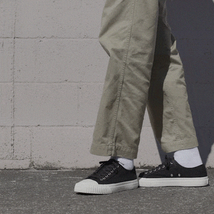 Person wearing black low-top sneakers and khaki pants shifting stance against a stone wall.
