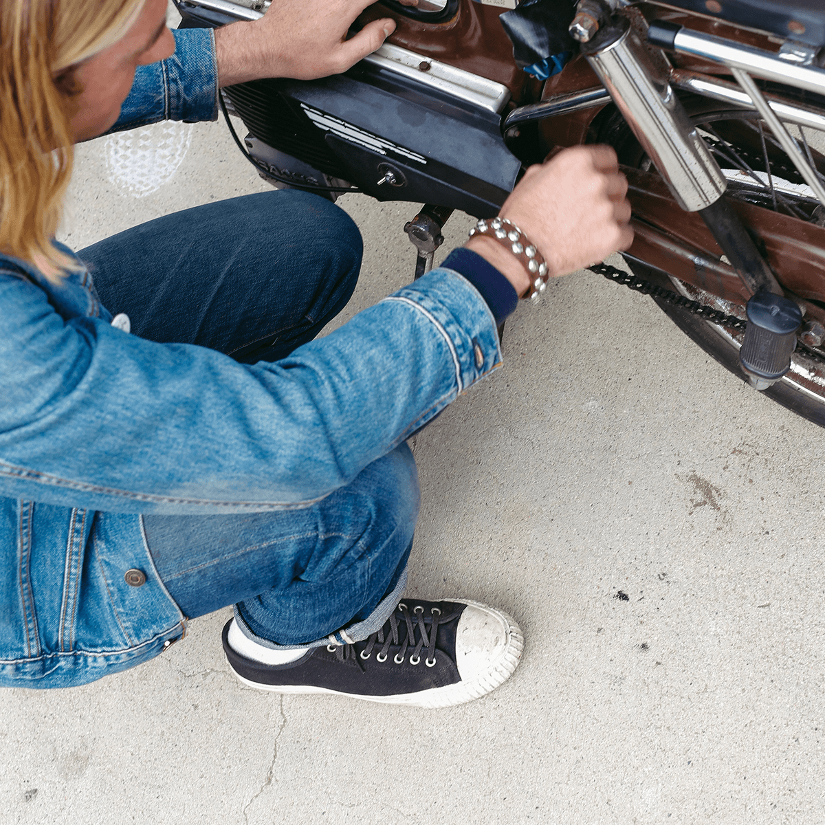 Man crouched while inspecting a motorcycle, wearing black low-top sneakers.
