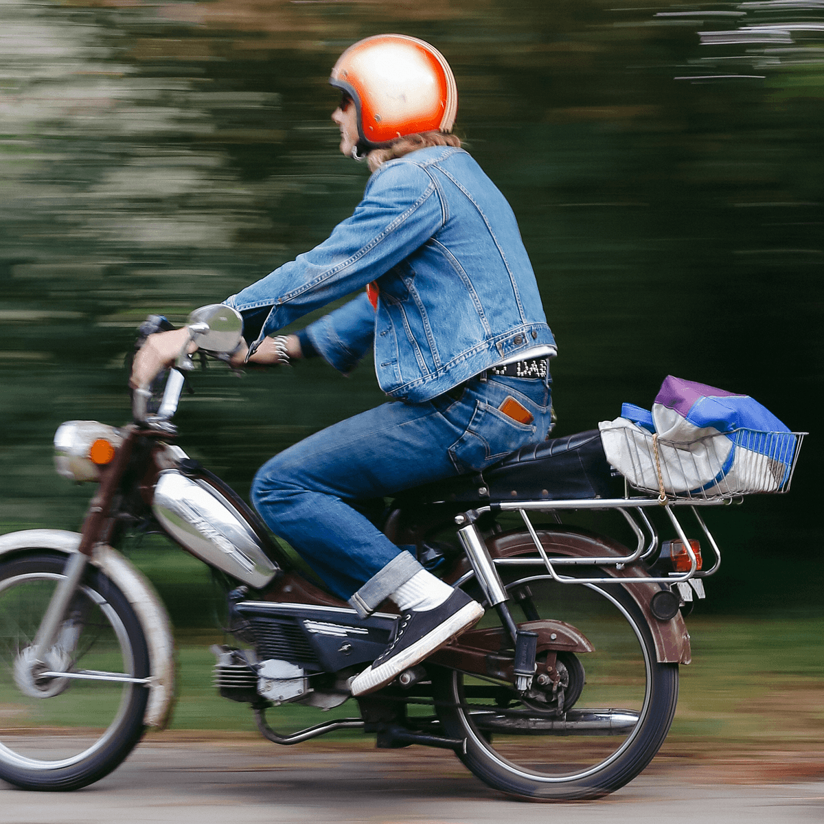 Man in a denim outfit and black low-top sneakers riding a motorcycle, with motion blur in the background.