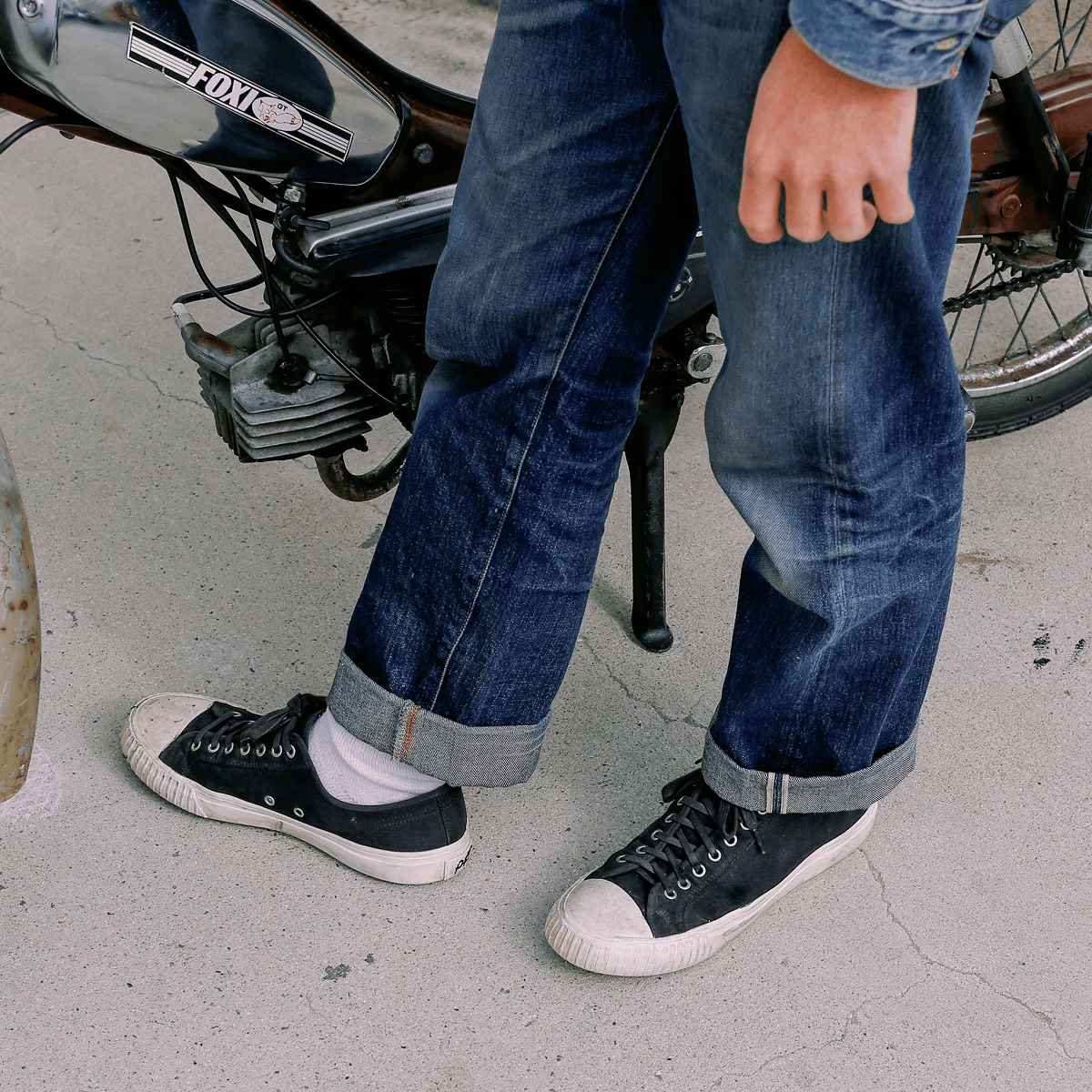 Person wearing blue jeans and black low-top sneakers standing next to a motorcycle.