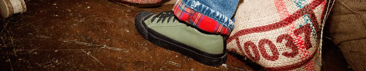 Olive green low-top sneaker with black midsoles worn on a distressed floor.