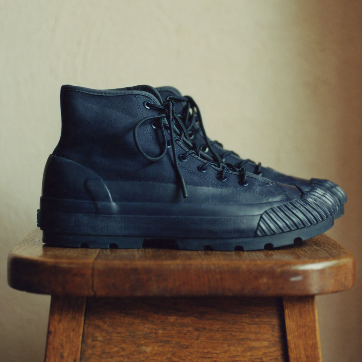 A pair of dark navy boots with lugged rubber outsoles and round hiker laces, resting on a wooden stool against a beige wall.
