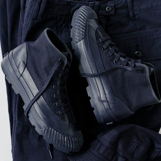 A pair of navy boots with lugged rubber outsoles, displayed on top of navy pants against a light gray background.