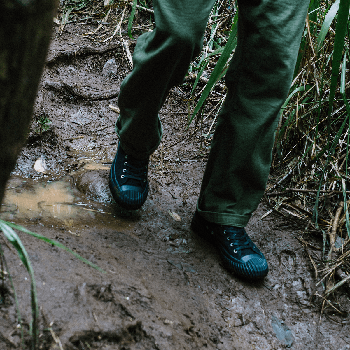 Person wearing green pants and black high-top sneakers walking along a muddy path.