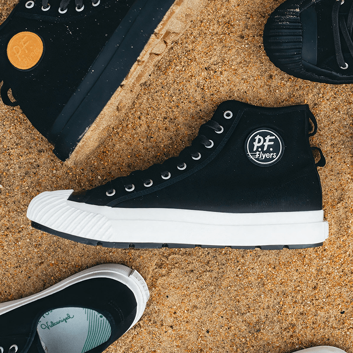 Close-up of a black high-top sneaker with white soles, surrounded by other sneakers on sand.