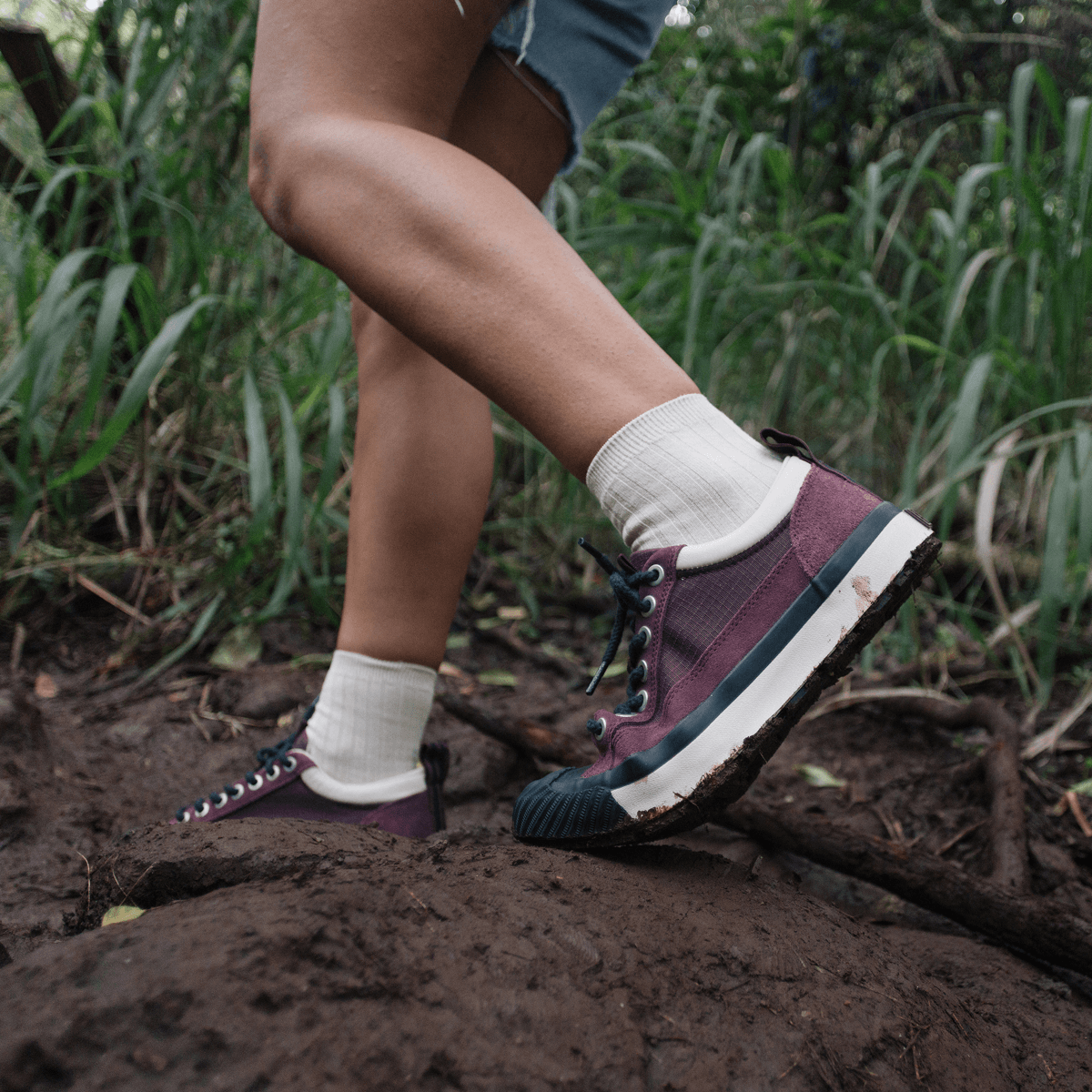 Person wearing burgundy low-top sneakers on a muddy trail with greenery in the background.