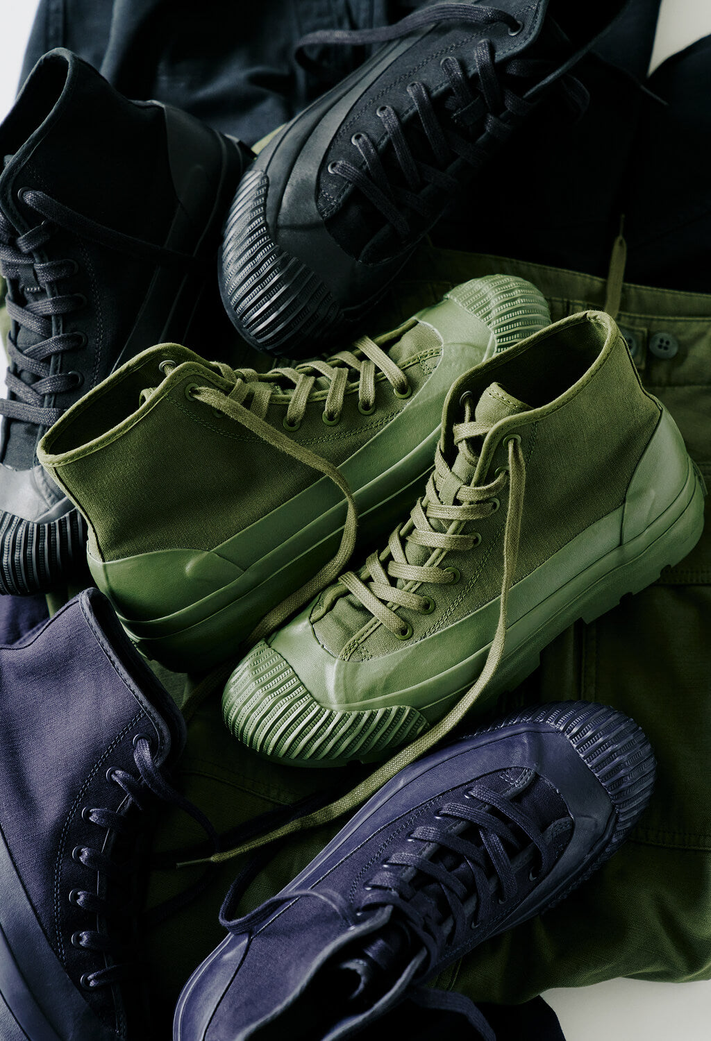 Three pairs of canvas boots in black, olive green, and navy, arranged on fabric against a light gray background.