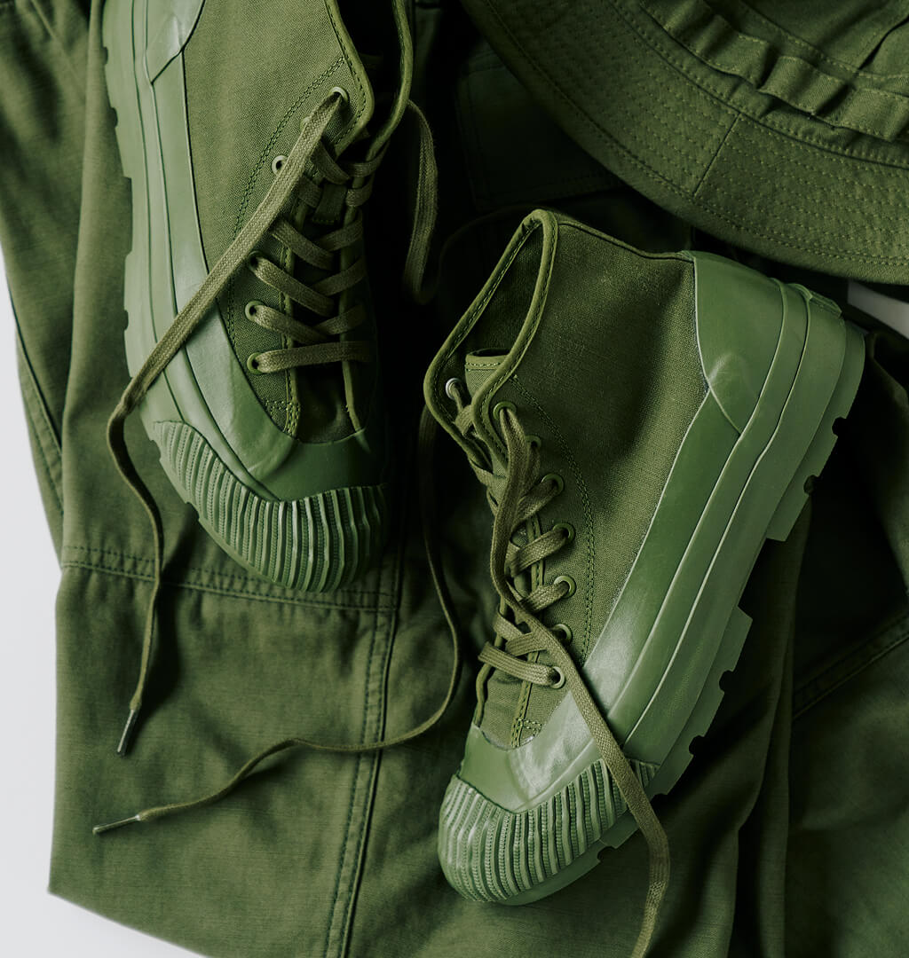 A pair of olive green boots with lugged rubber outsoles, displayed on top of an olive green pants against a light gray background.