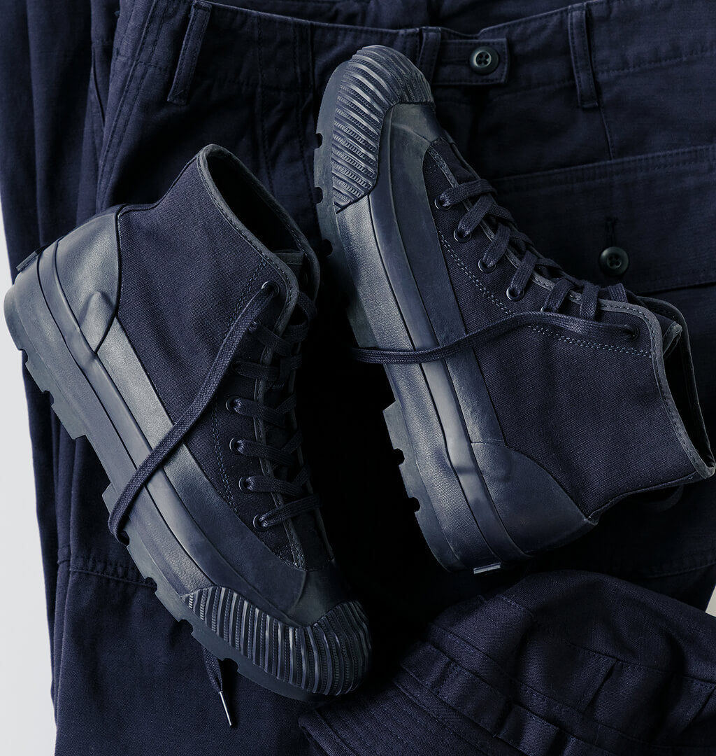 A pair of navy boots with lugged rubber outsoles, displayed on top of navy pants against a light gray background.