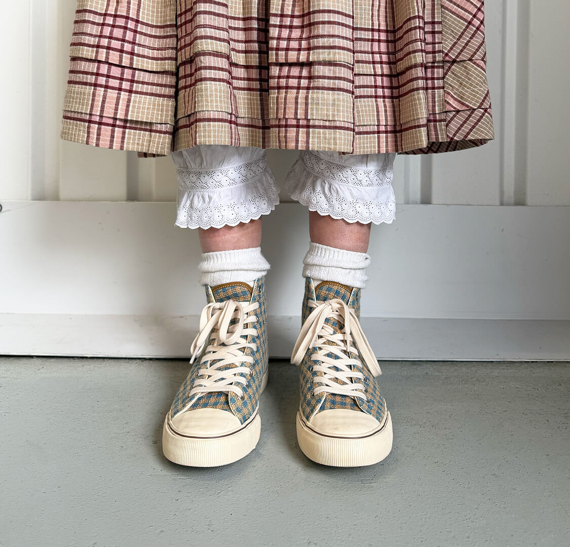 A person standing against a white paneled wall wearing indigo and brown plaid high-top sneakers.