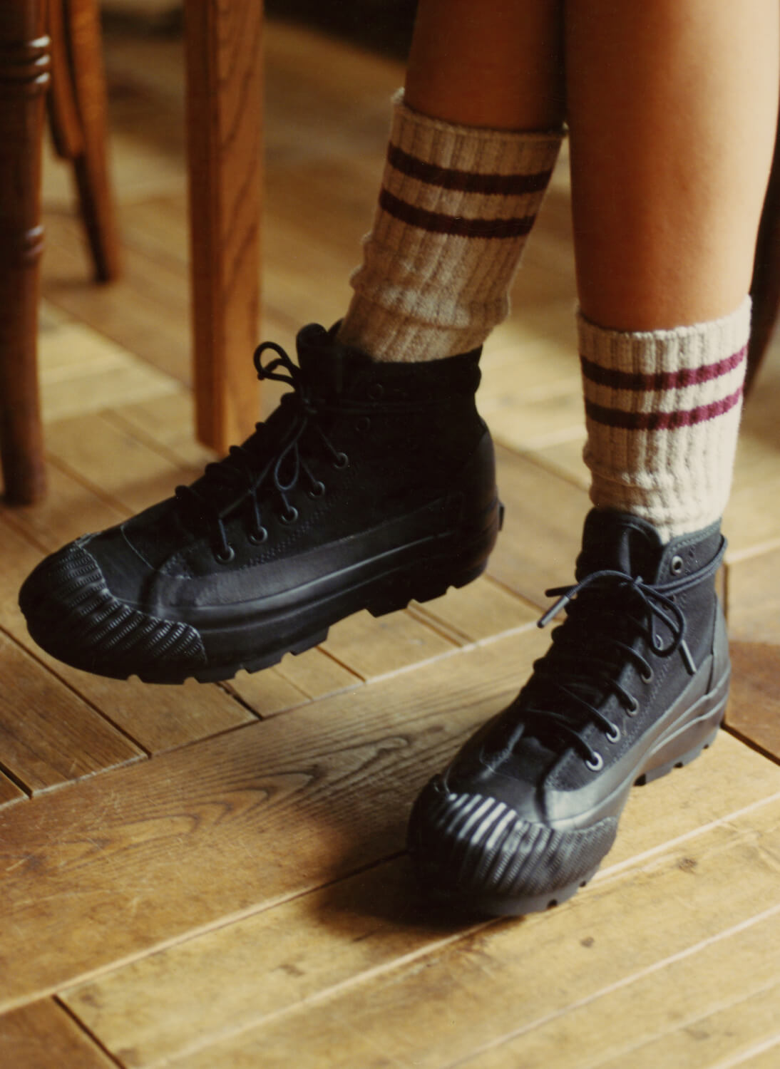 A person wearing black canvas boots with lugged rubber outsoles on a wooden floor.
