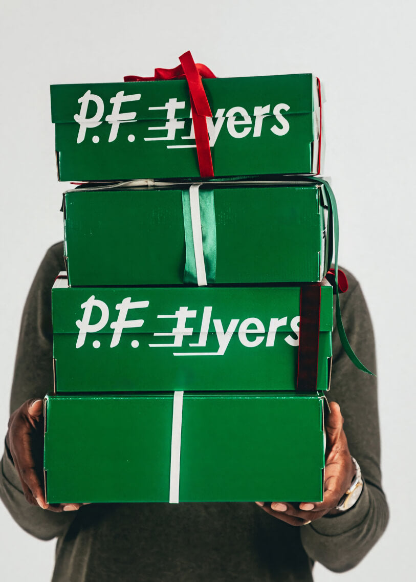 Stack of green P.F. Flyers shoe boxes held by a person against a light gray background