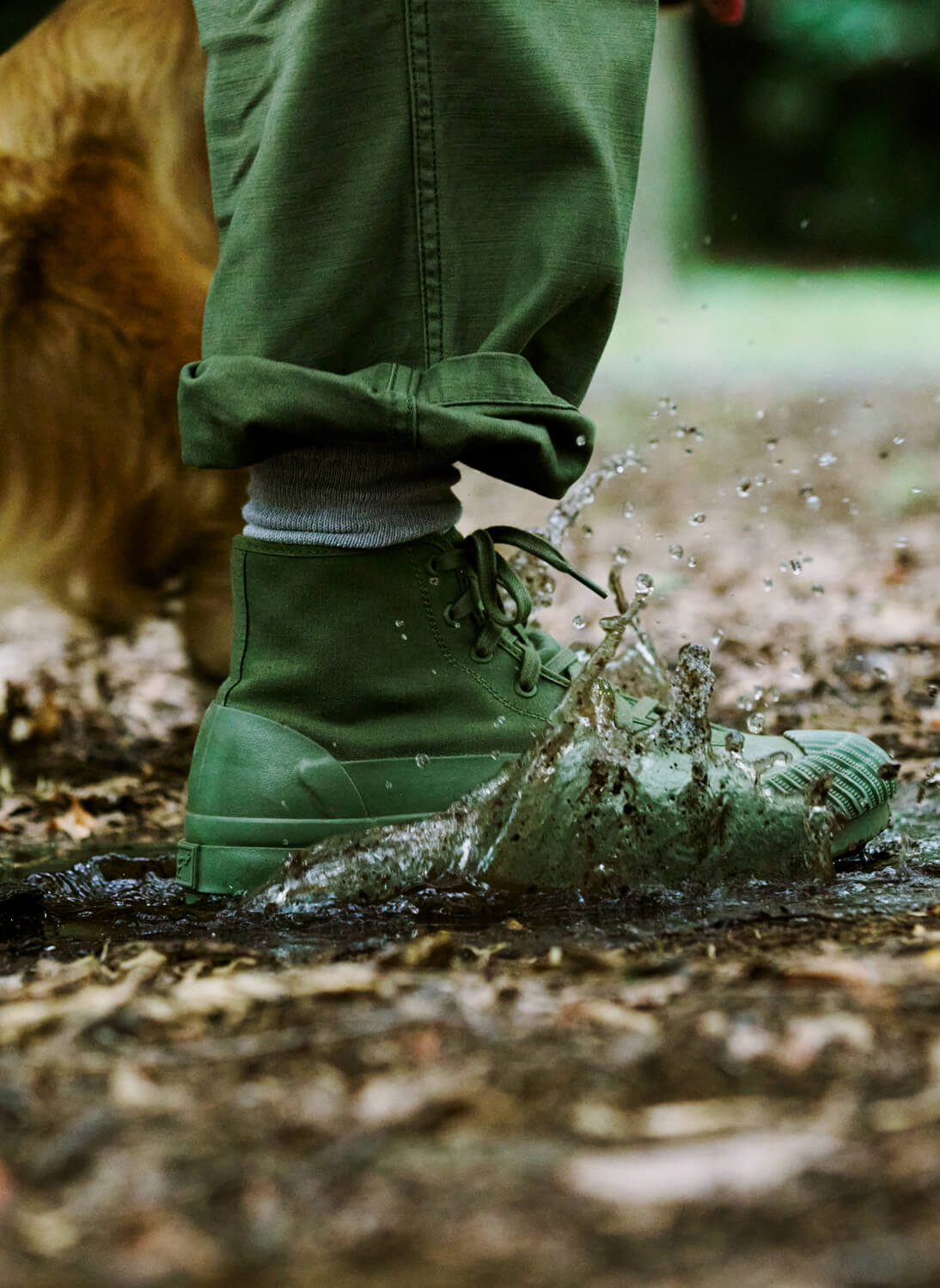 Close-up of a person wearing an olive green boot, stepping into a muddy puddle on a forest floor.
