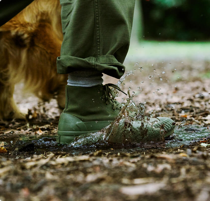 Close-up of a person wearing an olive green boot, stepping into a muddy puddle on a forest floor.