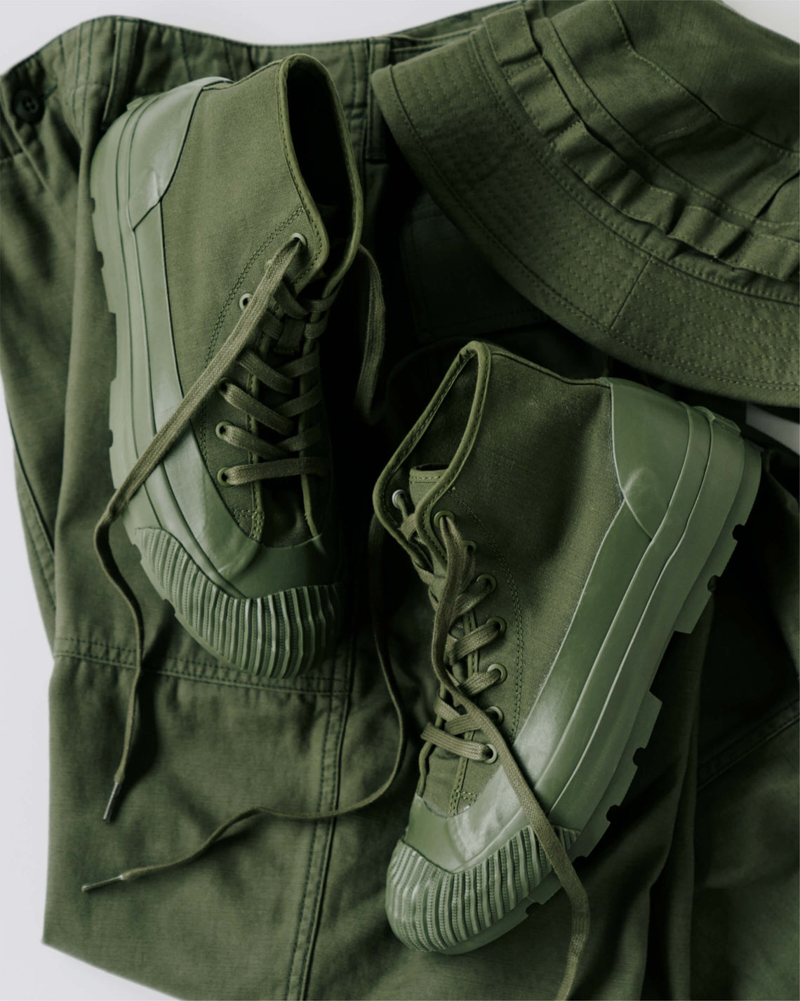 A pair of olive green boots with lugged rubber outsoles, displayed on top of an olive green pants against a light gray background.